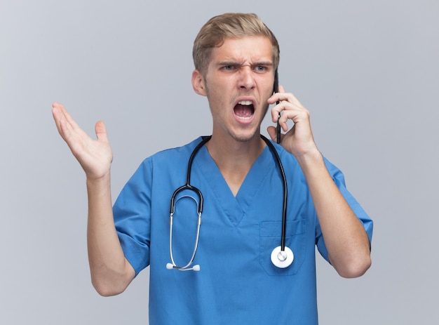 Il giovane medico maschio arrabbiato che porta l'uniforme del medico con lo stetoscopio parla sul telefono diffondendo la mano isolata sul muro bianco