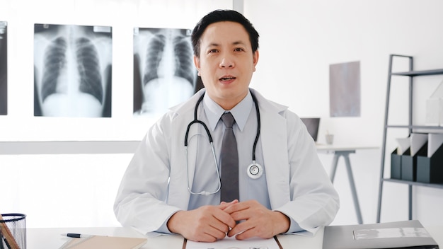 Il giovane medico maschio dell'Asia in uniforme medica bianca con lo stetoscopio utilizzando il computer portatile parla in videoconferenza con il paziente, guardando la telecamera nell'ospedale sanitario.