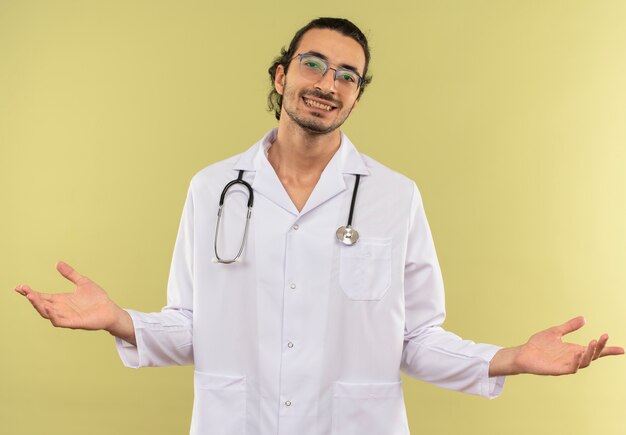 Il giovane medico maschio sorridente con i vetri ottici che indossa la veste bianca con lo stetoscopio diffonde le mani sulla parete verde isolata con lo spazio della copia