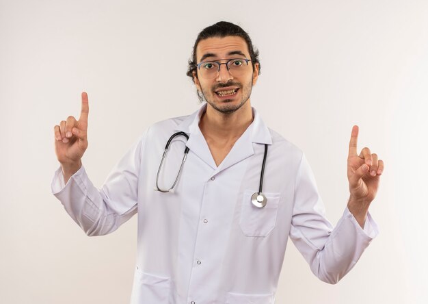 Il giovane medico maschio sorridente con i vetri ottici che indossa la veste bianca con lo stetoscopio indica fino alla parete bianca isolata con lo spazio della copia