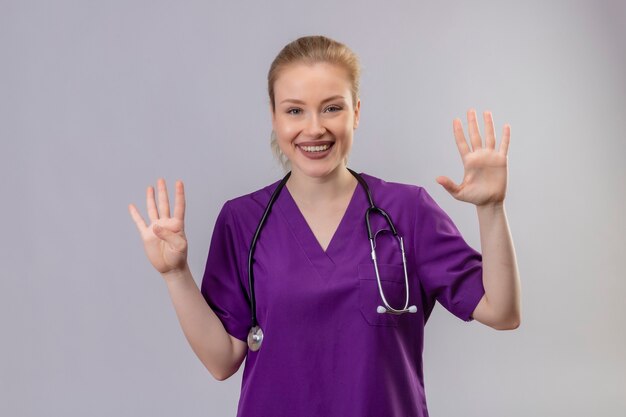 Il giovane medico sorridente che porta l'abito medico viola e lo stetoscopio mostra i numeri differenti sulla parete bianca isolata