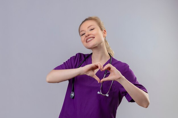 Il giovane medico sorridente che porta l'abito medico viola e lo stetoscopio mostra il gesto del cuore sulla parete bianca isolata