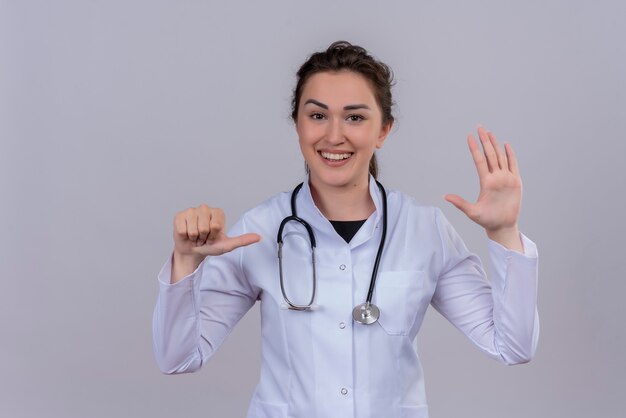 Il giovane medico sorridente che porta lo stetoscopio d'uso dell'abito medico mostra il gesto differente sulla parete bianca