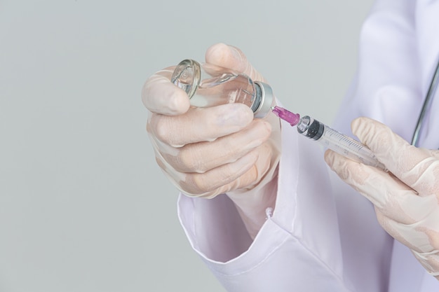 Il giovane medico sta tenendo la siringa ipodermica con i guanti di gomma della fiala del vaccino sulla parete grigia.