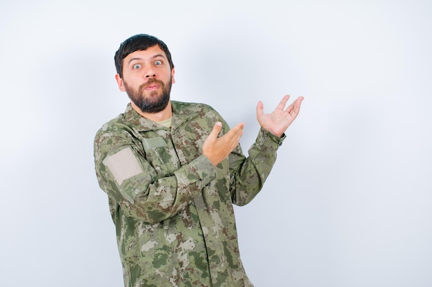 Il giovane militare sorpreso sta guardando la fotocamera puntando verso l'alto con le mani su sfondo bianco