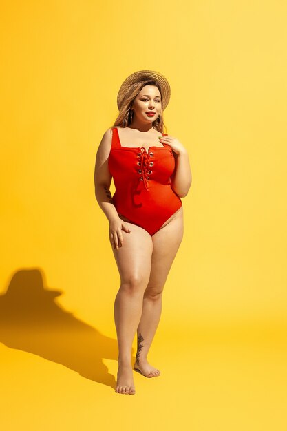 Il giovane modello femminile caucasico plus size si sta preparando per il resort sulla spiaggia sulla parete gialla. Donna in costume da bagno rosso e cappello in posa fiducioso. Concetto di estate, festa, corpo positivo, uguaglianza.