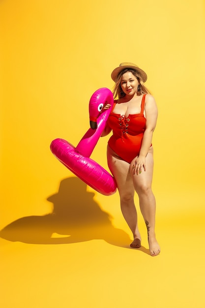 Il giovane modello femminile caucasico plus size sta preparando le vacanze sulla parete gialla. Donna in costume da bagno rosso e cappello in posa con swimring.