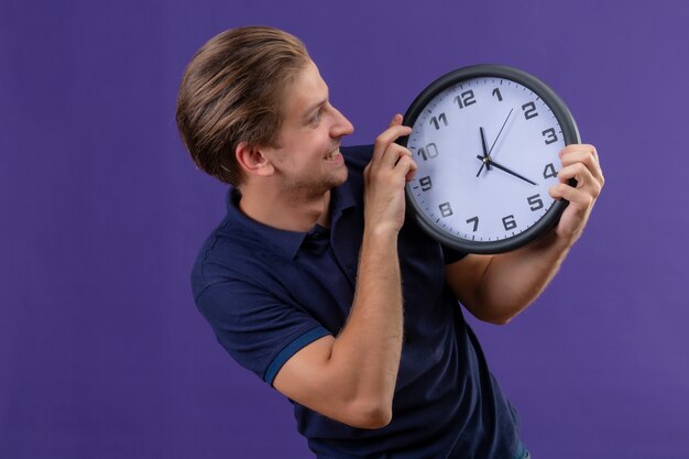 Il giovane orologio bello della tenuta del tipo in mani che lo esaminano con il fronte felice è uscito e allegro che controlla il fondo porpora