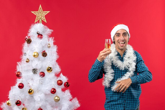 Il giovane pazzo con il cappello di Babbo Natale e alzando un bicchiere di vino si rallegra vicino all'albero di Natale
