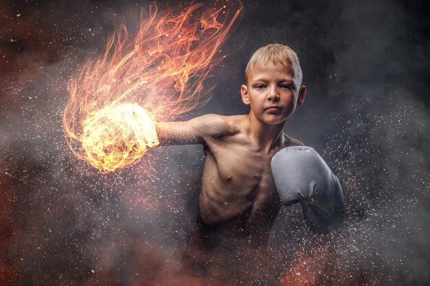 Il giovane pugile a torso nudo con i capelli biondi che indossa i guanti da boxe mostra un gancio da boxe. Isolato su uno sfondo scuro.