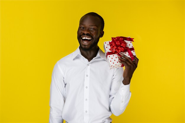 Il giovane ragazzo afroamericano barbuto sorridente emozionante sta tenendo un regalo nella mano sinistra e guarda davanti lui in camicia bianca