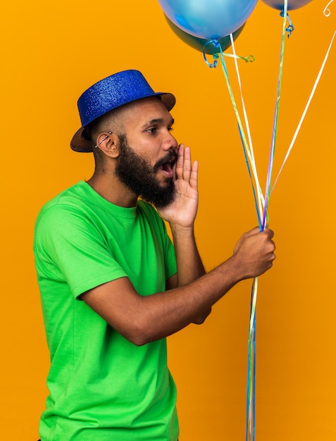 Il giovane ragazzo afroamericano sospettoso che indossa un cappello da festa tiene in mano palloncini sussurri isolati sul muro arancione