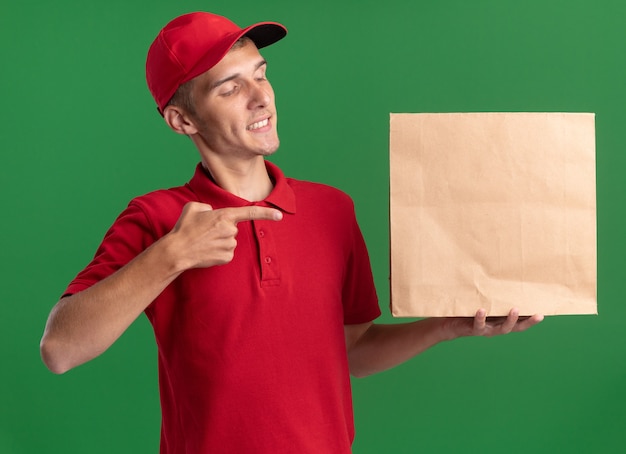 Il giovane ragazzo biondo di consegna soddisfatto osserva e indica il pacchetto di carta sul verde