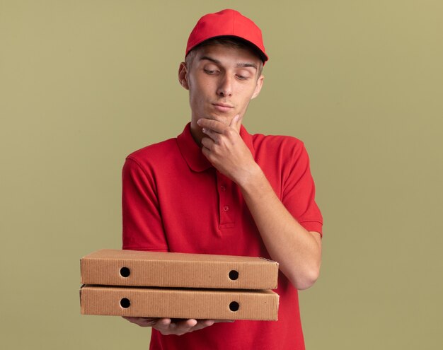 Il giovane ragazzo biondo e soddisfatto delle consegne mette la mano sul mento che tiene e guardando le scatole per pizza su verde oliva