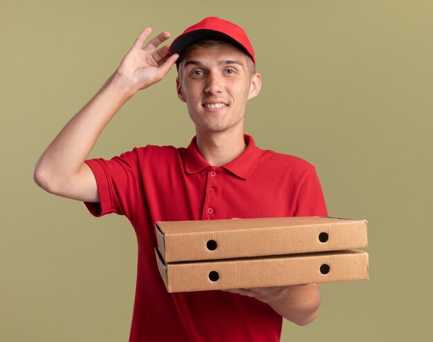 Il giovane ragazzo biondo sorridente delle consegne mette la mano sul cappuccio e tiene le scatole della pizza su verde oliva