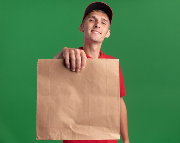 Il giovane ragazzo biondo sorridente delle consegne tiene il pacchetto di carta sul verde