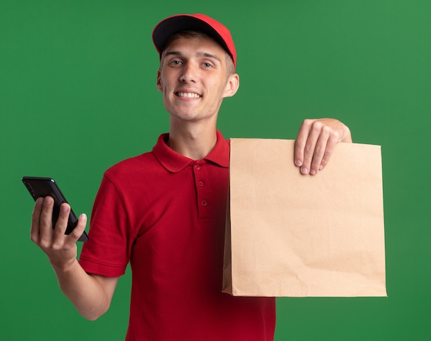 Il giovane ragazzo biondo sorridente delle consegne tiene il pacchetto e il telefono di carta sul verde