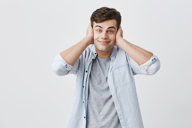 Il giovane ragazzo caucasico deridente triste si è vestito in camicia blu-chiaro, facendo i fronti, sorridendo, chiudendo le orecchie con le palme, rifiutando di ascoltare le parole del `s di qualcuno, posando.