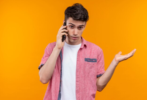 Il giovane ragazzo caucasico sorpreso che porta la camicia rosa parla sul telefono alzato la mano su fondo arancio isolato