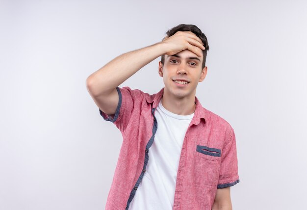 Il giovane ragazzo caucasico sorridente che porta la camicia rosa ha messo la sua mano sulla fronte su fondo bianco isolato