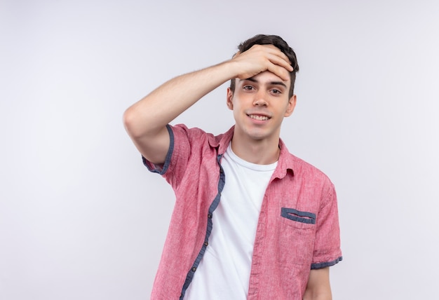 Il giovane ragazzo caucasico sorridente che porta la camicia rosa ha messo la sua mano sulla fronte su fondo bianco isolato