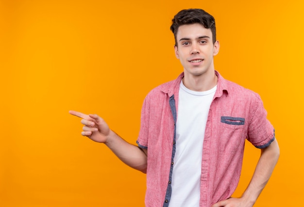 Il giovane ragazzo caucasico sorridente che porta la camicia rosa indica al lato su fondo arancio isolato