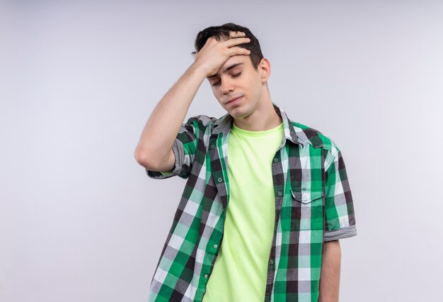 Il giovane ragazzo caucasico stanco che porta la camicia verde ha messo la sua mano sulla fronte su bianco isolato