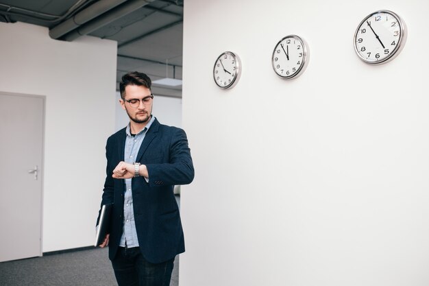 Il giovane ragazzo con gli occhiali sta camminando in ufficio. Indossa camicia blu, giacca scura, jeans e barba. Tiene il portatile in mano. Sta guardando l'orologio.