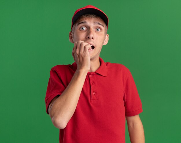 Il giovane ragazzo delle consegne biondo ansioso morde le unghie isolate sulla parete verde con lo spazio della copia