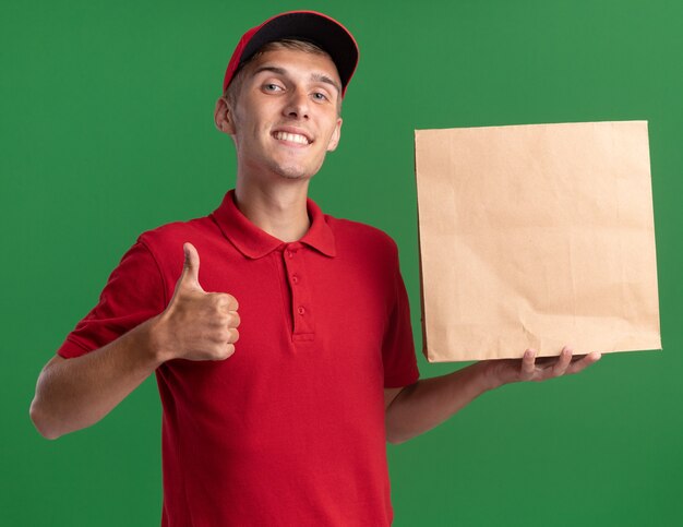 Il giovane ragazzo delle consegne biondo sorridente alza i pollici e tiene il pacchetto di carta isolato sulla parete verde con lo spazio della copia