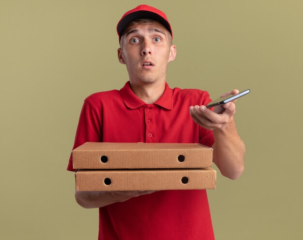 Il giovane ragazzo di consegna biondo ansioso tiene le scatole e il telefono della pizza isolati sulla parete verde oliva con lo spazio della copia