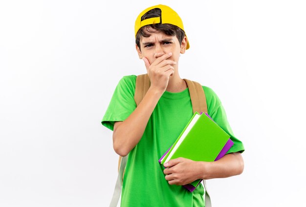 Il giovane ragazzo di scuola interessato che indossa lo zaino con il cappuccio che tiene i libri ha coperto la bocca con la mano isolata sulla parete bianca