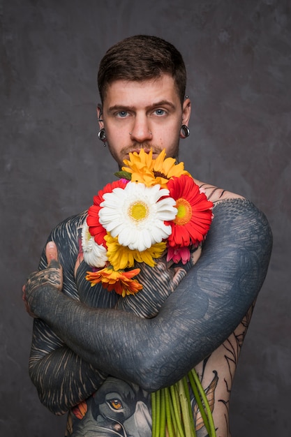 Il giovane senza camicia con tatuato sul suo corpo che tiene la bella gerbera fiorisce a disposizione che sta contro il fondo grigio