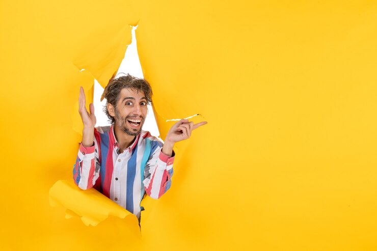 Foto gratuito il giovane sorridente felice ed emotivo posa sullo sfondo di un buco di carta gialla strappata