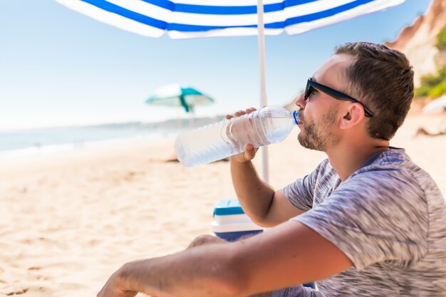 Il giovane sotto l'ombrello solare verde beve l'acqua sulla spiaggia?