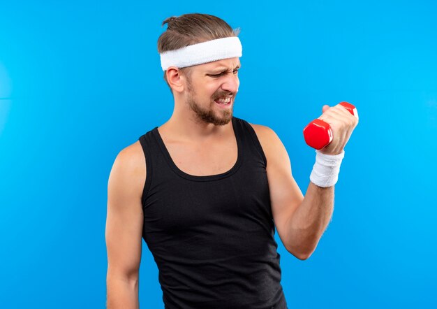 Il giovane sportivo bello dispiaciuto indossa la fascia e i braccialetti che tengono e che esaminano il dumbbell isolato sullo spazio blu