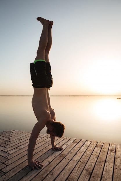 Il giovane sportivo bello fa gli esercizi di yoga alla spiaggia.