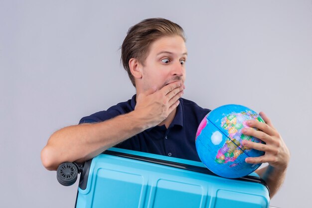 Il giovane tipo bello del viaggiatore che sta con il globo della tenuta della valigia che lo esamina stupito e sorpreso che copre la bocca con consegna il fondo bianco