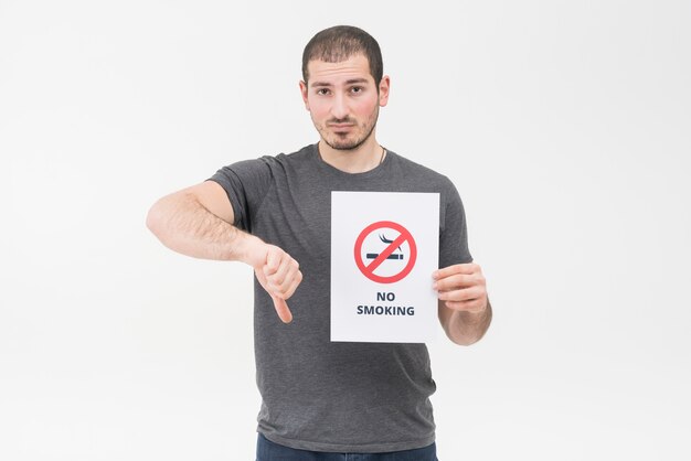 Il giovane triste che tiene il segno non fumatori che mostra il pollice giù gesture contro fondo bianco
