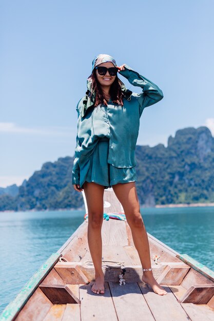 Il giovane turista felice asiatico del blogger della donna in vestito di seta e sciarpa e occhiali da sole in vacanza viaggia intorno alla Tailandia sulla barca asiatica, parco nazionale di Khao Sok.