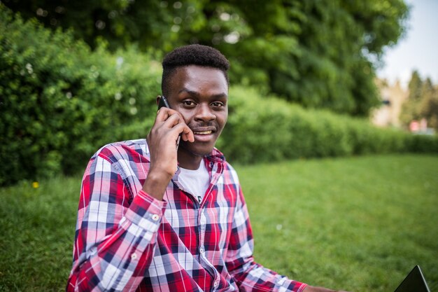 Il giovane uomo afroamericano parla al telefono mentre era seduto sull'erba verde con il computer portatile