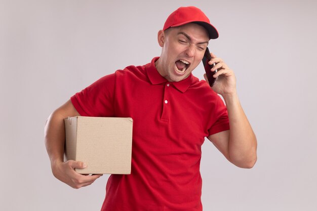 Il giovane uomo arrabbiato di consegna che porta l'uniforme con la scatola della tenuta del cappuccio parla sul telefono isolato sulla parete bianca