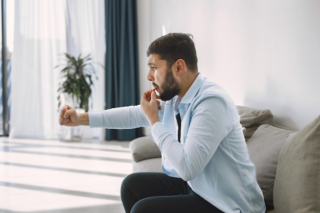 Il giovane uomo barbuto moderno eccitato bello si sta rilassando sul divano mentre guarda la partita di calcio in TV. L'uomo fischietta. Uomo in camicia blu seduto sul divano e tenendo le mani in aria.