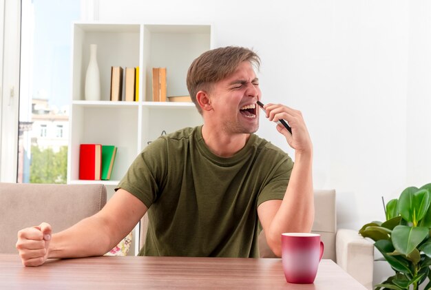 Il giovane uomo bello biondo dispiaciuto si siede al tavolo con la tazza mantenendo il pugno e tenendo il telefono