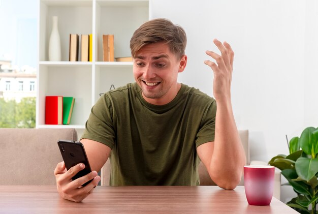 Il giovane uomo bello biondo sorpreso si siede al tavolo con la tazza che alza la mano che tiene e guardando il telefono all'interno del soggiorno