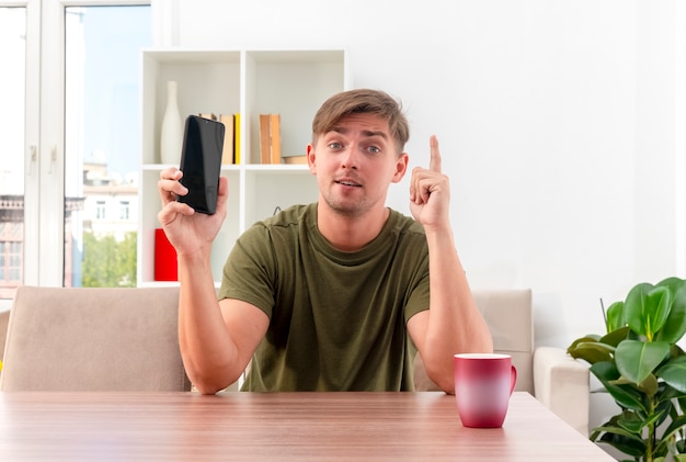 Il giovane uomo bello biondo sorpreso si siede alla tavola con il telefono della tenuta della tazza