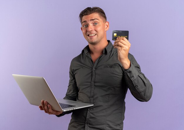 il giovane uomo bello biondo sorpreso tiene il computer portatile e la carta di credito