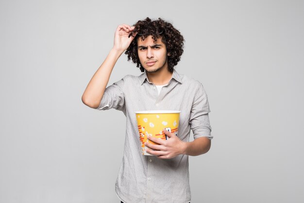 Il giovane uomo bello indossa i vetri 3d con capelli ricci che tengono una ciotola di popcorn sopra la parete bianca isolata
