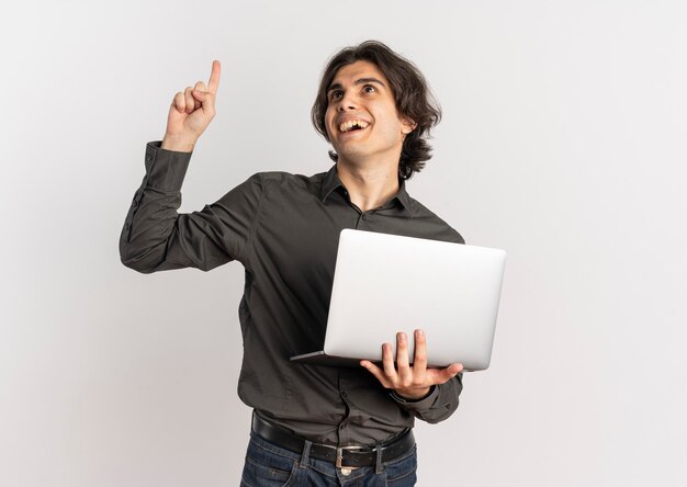Il giovane uomo caucasico bello sorpreso tiene il computer portatile e indica in alto isolato su priorità bassa bianca con lo spazio della copia