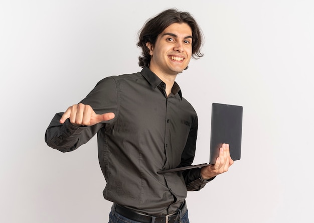 Il giovane uomo caucasico bello sorridente tiene e indica il computer portatile isolato su priorità bassa bianca con lo spazio della copia